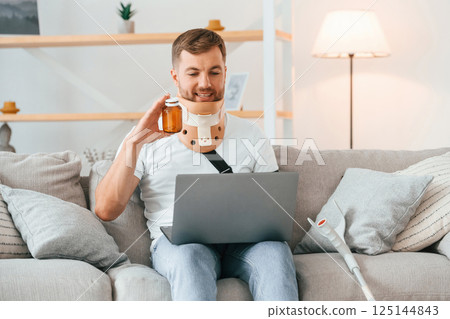 Holding pills, using laptop, having online conversation. Man with crutches is at home indoors. Having injuries 125144843