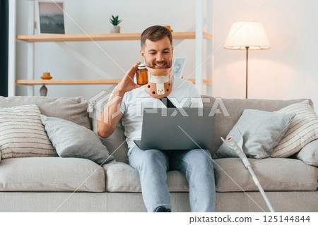 Holding pills, using laptop, having online conversation. Man with crutches is at home indoors. Having injuries 125144844