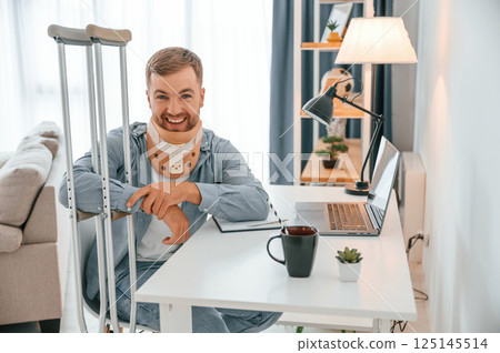 Sitting by table and smiling. Man with crutches is at home indoors. Having injuries 125145514