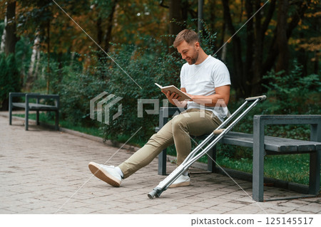 Sitting with the book. Man with crutches is in the park outdoors. Having leg injury 125145517