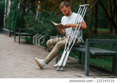 Sitting with the book. Man with crutches is in the park outdoors. Having leg injury 125145518