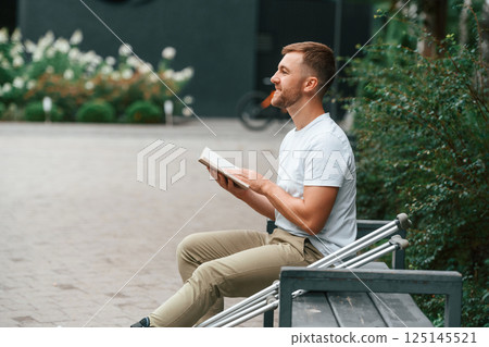 Sitting with the book. Man with crutches is in the park outdoors. Having leg injury 125145521