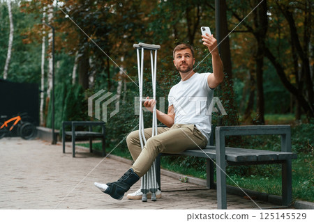 Using smartphone to make selfie. Man with crutches is in the park outdoors. Having leg injury 125145529