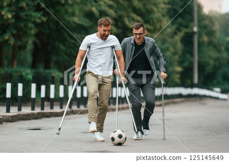 Playing with soccer ball. Two men with crutches is outdoors on the road 125145649