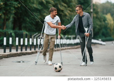Standing, having fun, playing soccer. Two men with crutches is outdoors on the road 125145653