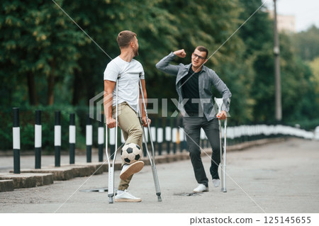 Standing, having fun, playing soccer. Two men with crutches is outdoors on the road 125145655