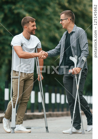 Doing handshake. Two men with crutches is outdoors on the road 125145662