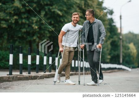 Standing and smiling. Two men with crutches is outdoors on the road 125145664