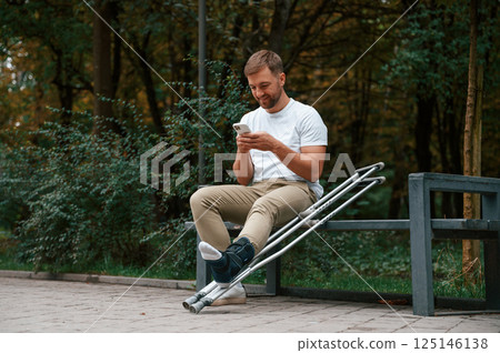 Sitting, holding smartphone. Man with crutches is in the park outdoors. Having leg injury 125146138