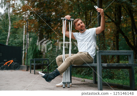Making a selfie. Man with crutches is in the park outdoors. Having leg injury 125146148