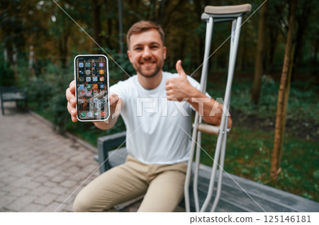 Holding and showing smartphone. Man with crutches is in the park outdoors. Having leg injury 125146181