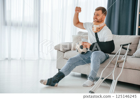 Sitting, holding soccer ball, watching sport match. Man with crutches is at home indoors. Having injuries 125146245