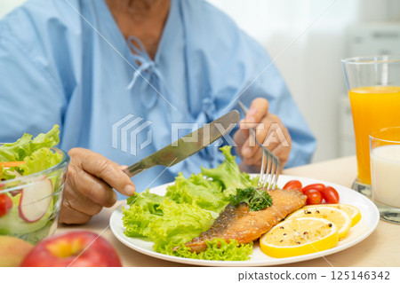 Asian senior woman patient eating Salmon steak breakfast with vegetable healthy food while sitting and hungry on bed in hospital. Asian senior woman patient eating Salmon steak breakfast with vegetable healthy food while sitting and hungry on bed in hospital. 125146342