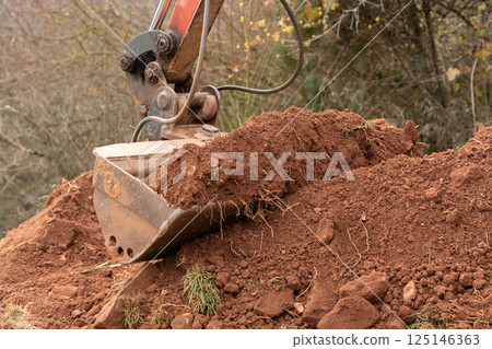 The excavator bucket filled with clay soil and stones, digging into the earth. The heavy machinery operates on a construction site, preparing the ground for further work 125146363