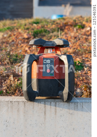 A laser level actively measuring on a land plot, ensuring precise alignment for construction and landscaping projects. The tool emits a red beam, aiding in accurate leveling on uneven terrain. A laser level actively measuring on a land plot, ensuring precise alignment for construction and landscaping projects. The tool emits a red beam, aiding in accurate leveling on uneven terrain. 125146391