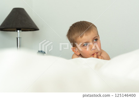 Child lying in a comfortable bed, appearing deep in thought. The curious expression on the child's face suggests contemplation or daydreaming. Warm, cozy indoor setting. High quality photo 125146400