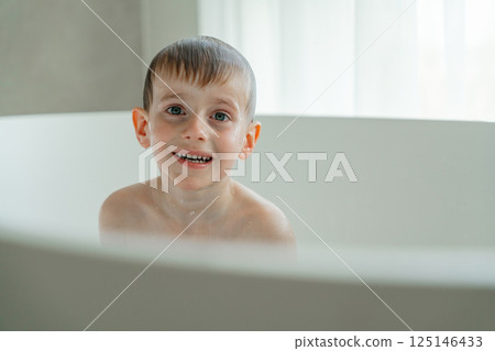 Young child happily enjoying bath time in a white tub, displaying a joyful expression with water droplets on skin. Bright atmosphere enhances the moment's playful mood. Young child happily enjoying bath time in a white tub, displaying a joyful expression with water droplets on skin. Bright atmosphere enhances the moment's playful mood. 125146433