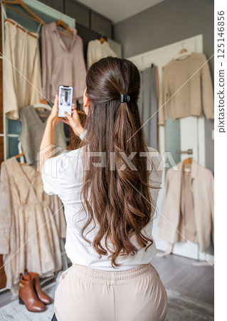 A young woman photographs clothing hanging on a wardrobe in bedroom using her smartphone. Online resale and second-hand fashion thrive through mobile apps, making sustainable shopping more accessible A young woman photographs clothing hanging on a wardrobe in bedroom using her smartphone. Online resale and second-hand fashion thrive through mobile apps, making sustainable shopping more accessible 125146856