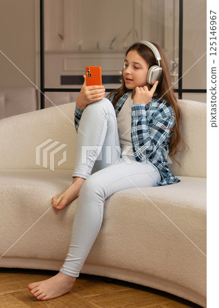 Girl sitting on sofa wearing headphones and using smartphone. 125146967