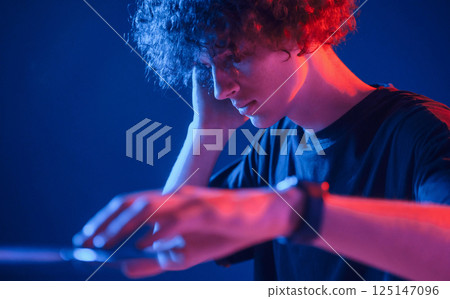 DJ is in the club. Young man with curly hair is indoors illuminated by neon lighting DJ is in the club. Young man with curly hair is indoors illuminated by neon lighting 125147096