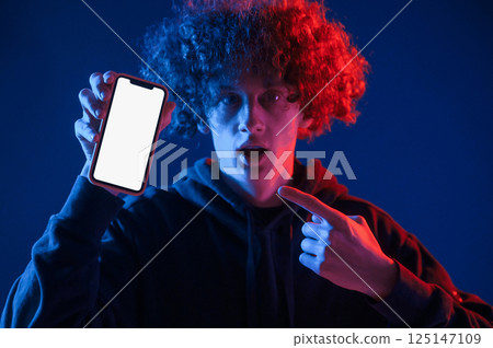 Holding smartphone and showing it to the camera. Young man with curly hair is indoors illuminated by neon lighting 125147109