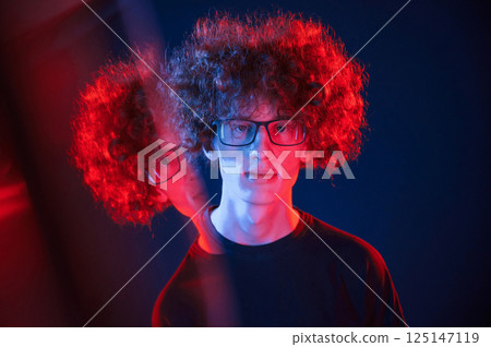 Glass reflection on the left side. Young man with curly hair is indoors illuminated by neon lighting 125147119
