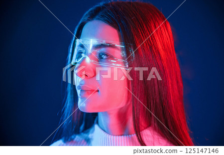 In futuristic eyewear. Young beautiful woman is indoors in the studio illuminated by neon light In futuristic eyewear. Young beautiful woman is indoors in the studio illuminated by neon light 125147146