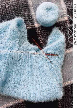 Blue cotton knitting with knitting needles on plaid. Soft blue knitwear with wooden circle needles. Knitting pattern background. Knitting lifestyle. Cozy hobby. Warm knitwear. Craft product concept 125148471