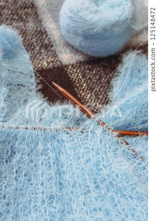 Blue cotton knitting with knitting needles on plaid. Soft blue knitwear with wooden circle needles. Knitting pattern background. Knitting lifestyle. Cozy hobby. Warm knitwear. Craft product concept 125148472