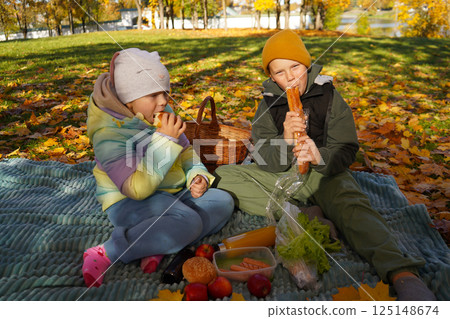 Wrapped in the warmth of friendship, a boy and a girl sit on a plaid in the fall park, sharing delicious food and enjoying the season. Wrapped in the warmth of friendship, a boy and a girl sit on a plaid in the fall park, sharing delicious food and enjoying the season. 125148674