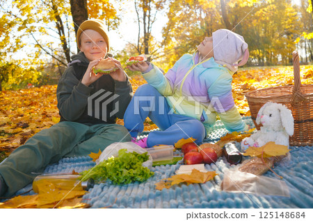 A boy and girl enjoy a delightful picnic in a public park on a sunny autumn day. Surrounded by vibrant fall foliage, they share snacks and laughter, embracing the warmth and beauty of the season. A boy and girl enjoy a delightful picnic in a public park on a sunny autumn day. Surrounded by vibrant fall foliage, they share snacks and laughter, embracing the warmth and beauty of the season. 125148684