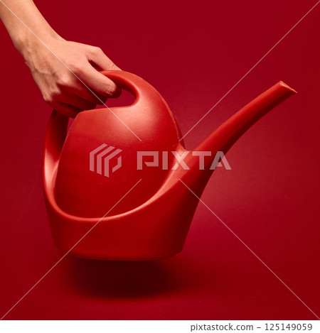 Hand gripping red watering can with elegant shape on red background. Concept of retro aesthetics, vintage, pop art, design Hand gripping red watering can with elegant shape on red background. Concept of retro aesthetics, vintage, pop art, design 125149059