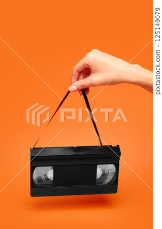Woman holding VHS tape by fragile magnetic ribbon like vintage handbag on orange backdrop. Woman holding VHS tape by fragile magnetic ribbon like vintage handbag on orange backdrop. 125149079