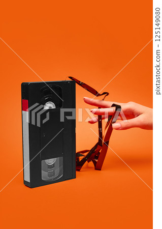 Hand with tangled VHS tape extending from cassette on orange background. Concept of retro aesthetics, vintage, pop art, design. 125149080