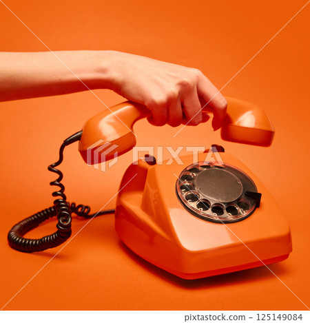 Hand lifts the receiver of a vintage orange rotary dial telephone against a matching orange backdrop. Hand lifts the receiver of a vintage orange rotary dial telephone against a matching orange backdrop. 125149084