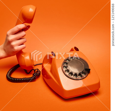 Hand lifting bright orange receiver, retro phone on orange background. Concept of human connection, urgent conversation, nostalgia. 125149088