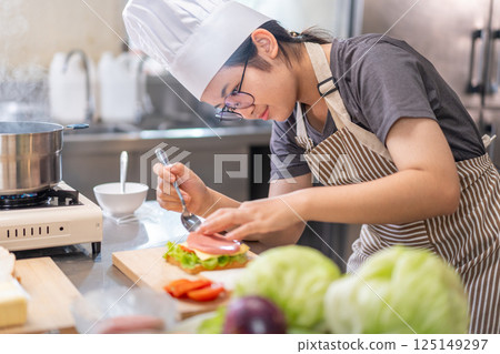 Smiling Young Female Chef Preparing a Sandwich in a Professional Kitchen Smiling Young Female Chef Preparing a Sandwich in a Professional Kitchen 125149297