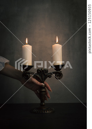 Female hand in white vintage dress sleeve take old brass candelabrum with burning candles from table in the dark, vertical image. Female hand in white vintage dress sleeve take old brass candelabrum with burning candles from table in the dark, vertical image. 125150935