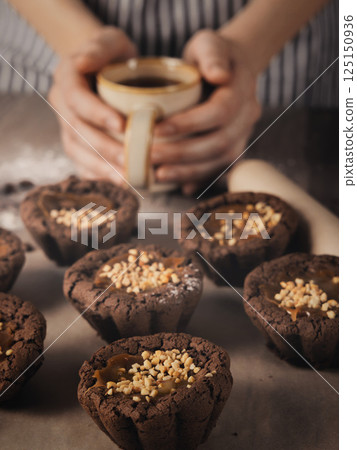 Female hands hold coffee mug and freshly baked muffins on baking paper on a table, vertical image. 125150936