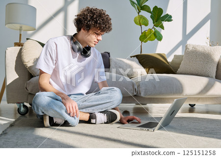 Handsome young male typing on laptop sitting on floor at home Handsome young male typing on laptop sitting on floor at home 125151258