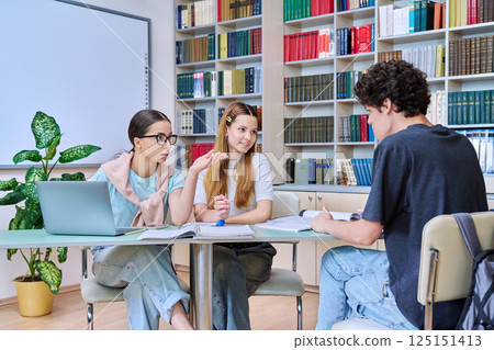 Group of students teenagers sitting together at desk in library preparing for tests exams Group of students teenagers sitting together at desk in library preparing for tests exams 125151413