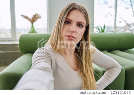 Selfie portrait serious confused beautiful young woman in home interior 125151655