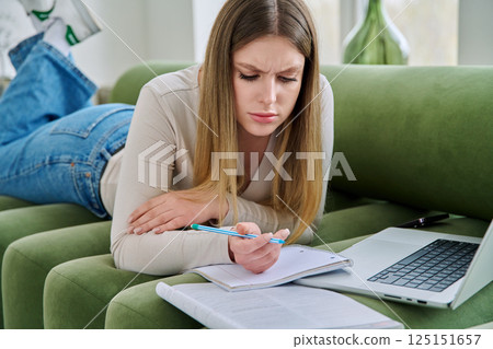 Young student girl lying at home on couch with laptop, notebook, textbook 125151657