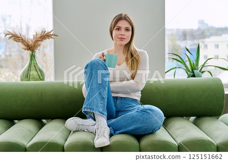 Portrait of relaxed beautiful young woman sitting at home on sofa with cup 125151662