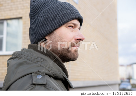 Handsome man wearing warm jacket and wool hat walking down on the city with mobile phone in hand. Stylish bearded middle-aged male using smartphone at urban street. Businessman going on job. 125152635