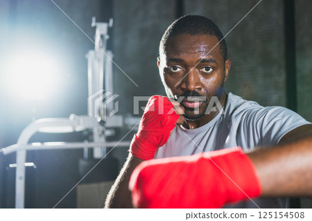 Workout in gym. African man fighter ready for fight punching with boxing wraps protective bandages to camera. Strong man training punches looking concentrated straight preparing for boxing sparring Workout in gym. African man fighter ready for fight punching with boxing wraps protective bandages to camera. Strong man training punches looking concentrated straight preparing for boxing sparring 125154508