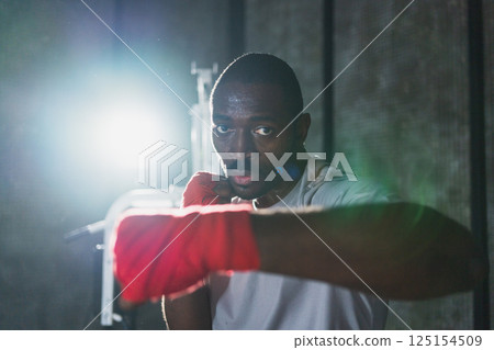 Workout in gym. African man fighter ready for fight punching with boxing wraps protective bandages to camera. Strong man training punches looking concentrated straight preparing for boxing sparring Workout in gym. African man fighter ready for fight punching with boxing wraps protective bandages to camera. Strong man training punches looking concentrated straight preparing for boxing sparring 125154509