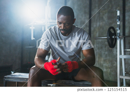 Fitness workout in gym. African American man fighter preparing for fight wrapping hands with red boxing wraps sports protective bandages in gym. Strong man ready for fight boxing sparring training 125154515