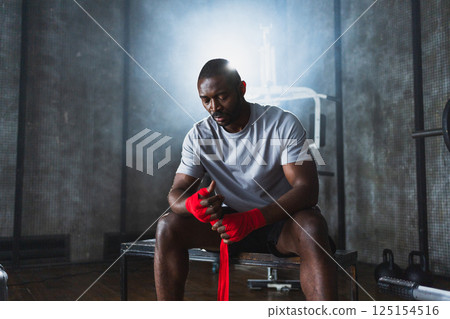 Fitness workout in gym. African American man fighter preparing for fight wrapping hands with red boxing wraps sports protective bandages in gym. Strong man ready for fight boxing sparring training 125154516