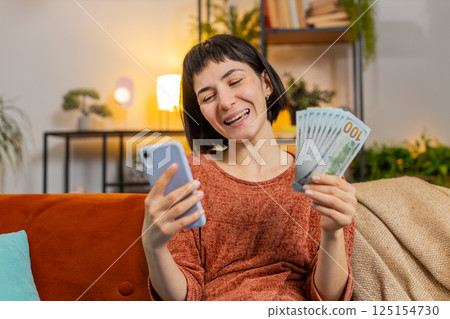 Woman counting money dollar banknotes savings earnings using smartphone calculator on sofa at home 125154730
